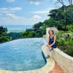 Woman sits on the edge of an infinity pool, with a tropical garden and distant ocean under a blue sky in the background.
